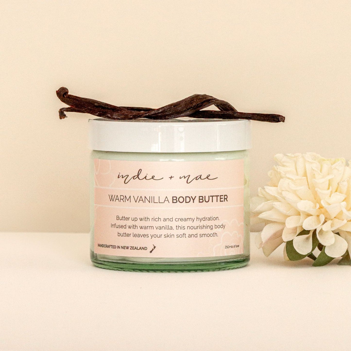 Jar of 'Indie + Mae Warm Vanilla Body Butter' with vanilla beans and flowers on a beige background