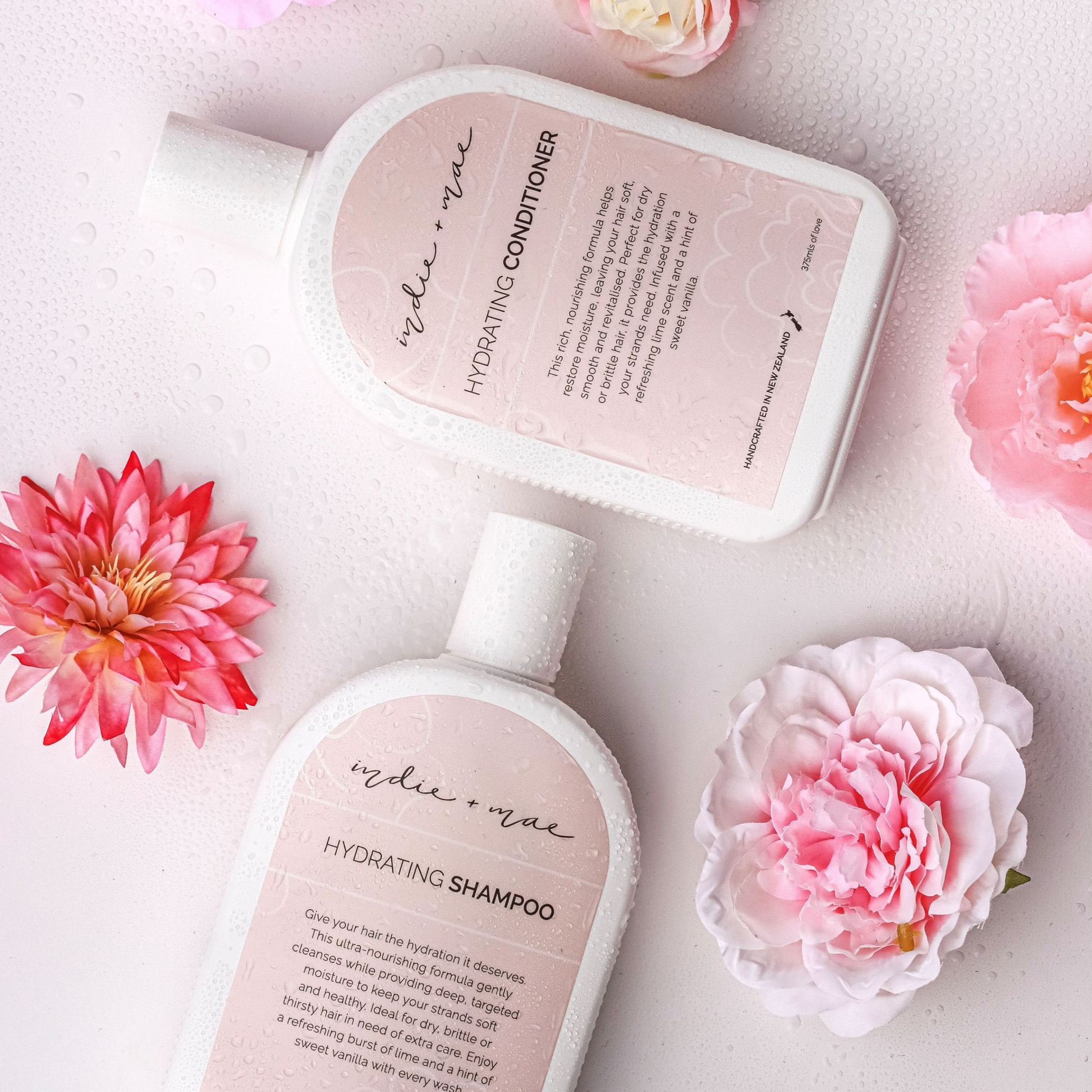 Two bottles of 'Indie + Mae' shampoo and conditioner with pink flowers on a light background