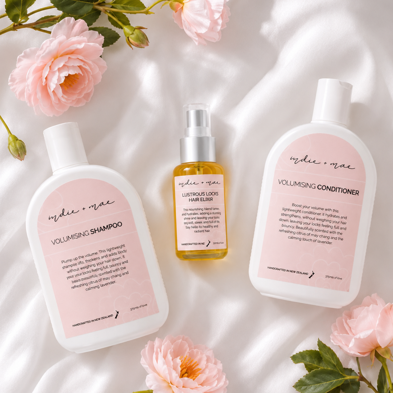 Three haircare products from the brand 'indie + mae' are arranged in a styled flat-lay on white draped fabric with pink roses. The set includes Volumising Shampoo, Volumising Conditioner, and Lustrous Locks Hair Elixir.